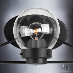 A close-up of the Urban Ambiance UHP2707 Contemporary Bathroom Vanity Light from the Aberdeen Collection, featuring a midnight black finish and a clear glass globe that reveals an illuminated filament bulb, is mounted on a gray wall.