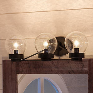 Three clear glass globe bath fixtures from the Urban Ambiance Aberdeen Collection are mounted on a midnight black wall plate above a wooden framed mirror, adding an industrial vanity light touch to the space with the UHP2707 Contemporary Bathroom Vanity Light (6.5″H x 22″W, Midnight Black Finish).