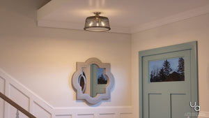 A UHP2732 Vintage Ceiling Fixture from Urban Ambiance’s Eugene Collection, featuring a brushed nickel finish and measuring 10.25 inches high by 16 inches wide, hangs from the ceiling above a decorative wall mirror and a blue door, with trees visible through the window.