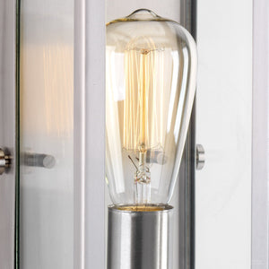 A clear vintage-style Edison light bulb with a visible filament is mounted in a brushed stainless steel fixture from the Messina Collection—the UHP2750 Luxe Industrial Electric Bath / Wall Light by Urban Ambiance—featuring a glass panel and sleek 12″H x 5-1/4″W stainless steel frame.