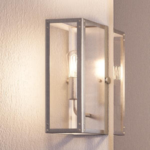 The Urban Ambiance UHP2750 Luxe Industrial Electric Bath / Wall Light from the Messina Collection, featuring a stainless steel finish and exposed bulb, is mounted against a textured light-colored wall. Measuring 12″ high by 5-1/4″ wide, this modern fixture blends sleek industrial design with contemporary sophistication.