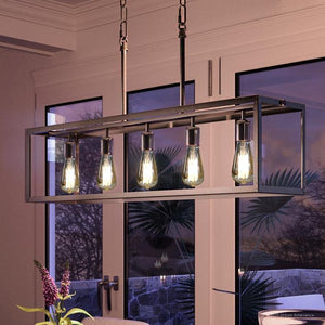 A sleek Urban Ambiance UHP2754 Modern Farmhouse Chandelier from the Messina Collection, measuring 9-3/4″ x 38″ with a stainless steel finish, hangs from the ceiling in a modern room with large glass doors revealing an evening outdoor view.