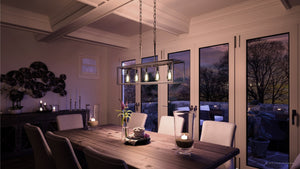 A modern dining room featuring a wooden table, eight chairs, and an Urban Ambiance UHP2754 Modern Farmhouse Chandelier from the Messina Collection in stainless steel finish, complemented by candles and glass doors revealing a twilight outdoor view.