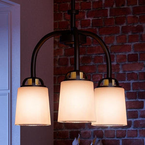 The UHP2764 Transitional Chandelier from Urban Ambiance’s Boise Collection, featuring an Olde Bronze finish and frosted glass shades, hangs from the ceiling in a room with exposed brick walls.