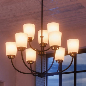 A lit Urban Ambiance UHP2768 Transitional Chandelier from the Boise Collection, featuring a sophisticated Olde Bronze finish and nine frosted glass lampshades, hangs elegantly from the ceiling in a room with a window revealing an evening sky—a striking showcase of Boise lighting fixtures.