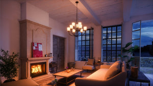 A cozy living room with a lit fireplace, beige sofas, a coffee table, indoor plants, large windows, and an elegant Urban Ambiance UHP2768 Transitional Chandelier from the Boise Collection in Olde Bronze finish—measuring 20-3/8″ x 31-1/4″—illuminates the space as it overlooks an evening outdoor view.