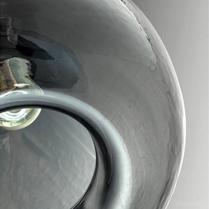 Close-up of the Urban Ambiance UHP2770 Modern Farmhouse Pendant from the Hobart Collection, featuring a glossy, reflective circular design with a hollow center, crafted from textured glass in a charcoal finish, set against a plain gray background.