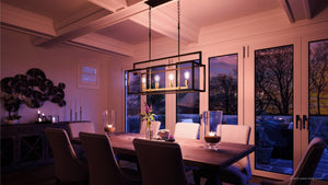 A modern dining room featuring a rectangular wooden table, cushioned chairs, candles, and the Urban Ambiance UHP2801 Modern Farmhouse Chandelier from the Cordoba Collection (17-1/4″ x 40-7/8″, Charcoal Finish), beautifully illuminated against large windows showcasing a sunset view.