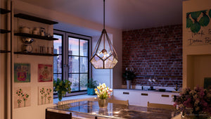 Modern kitchen with exposed brick walls, large windows revealing a cityscape at sunset, white cabinets, flowers on the table, and a striking Urban Ambiance UHP2851 Mid-Century Modern Chandelier from the Bari Collection in Olde Bronze finish (25-7/8″ x 19″) hanging above.