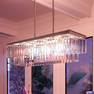 An Urban Ambiance UHP2883 Cosmopolitan Chandelier from the Lille Collection, measuring 11″ by 33-7/8″ and featuring an Antique Silver finish, hangs from the ceiling. Its prismatic glass is beautifully illuminated with a warm light above a window that frames a view of trees and plants outside.