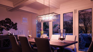 Modern dining room with a long wooden table, upholstered chairs, and candles, illuminated by the Urban Ambiance UHP2883 Cosmopolitan Chandelier (11″ x 33-7/8″) from the Lille Collection in an Antique Silver finish. Large windows reveal a sunset view framed by trees outside, while the chandelier’s prismatic glass from Urban Ambiance beautifully catches the evening light.