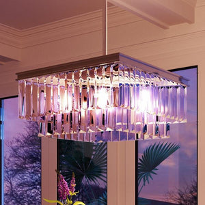 The Urban Ambiance UHP2885 Cosmopolitan Chandelier from the Lille Collection, measuring 11″ x 20-1/8″ with an antique silver finish, features a rectangular prismatic design adorned with multiple hanging clear glass prisms. Suspended from the ceiling in front of expansive windows at sunset and illuminated with a purple glow, it serves as an exquisite example of luxurious decorative lighting.