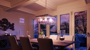 Contemporary dining room featuring a wooden table with eight chairs, glass candle holders, and luxurious decorative lighting highlighted by the UHP2885 Cosmopolitan Chandelier (11″ x 20-1/8″, Antique Silver Finish) from Urban Ambiance’s Lille Collection. Large windows frame views of a sunset and trees outside.