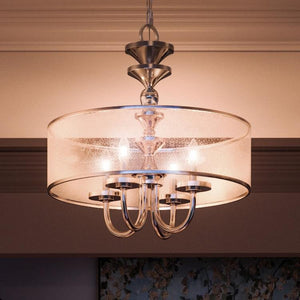 The Urban Ambiance UHP2922 Cosmopolitan Chandelier from the Charnwood Collection, featuring a polished nickel finish and measuring 20″ x 22″, hangs from the ceiling in a softly lit room, showcasing its modern, vintage-inspired design with four lights, metal arms, and a round frosted glass shade.