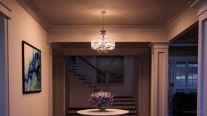 A hallway features a round table topped with a vase of purple flowers, illuminated by the Urban Ambiance UHP2922 Cosmopolitan Chandelier (20″ x 22″, Polished Nickel Finish) from the Charnwood Collection, with stairs in the background and two paintings adorning the walls.
