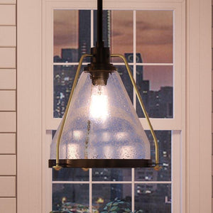 The UHP2991 Industrial Pendant Light from the Rochdale Collection by Urban Ambiance, featuring a 13″ x 13″ seeded glass shade and Olde Bronze finish, hangs in front of a window revealing a city skyline at dusk.