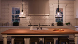 A modern kitchen features a white tile backsplash and a wooden island with stools, illuminated by two Urban Ambiance UHP2991 Industrial Pendant Lights (13″ x 13″, Olde Bronze Finish, Rochdale Collection), which cast a warm glow over the space. On the island sits a vase with branches and a cutting board with bread, while two windows reveal a night cityscape in the background.