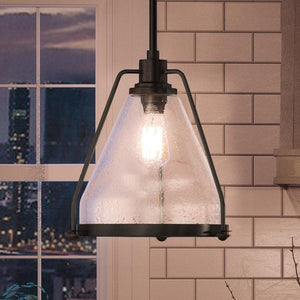The Urban Ambiance UHP2993 Industrial Pendant Light from the Rochdale Collection, featuring a clear geometric design and charcoal finish, hangs in front of a tiled wall and a window overlooking a cityscape at dusk, embodying an industrial-chic aesthetic.