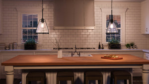 Modern kitchen with white cabinets, tiled backsplash, a wooden island, an Urban Ambiance UHP2993 Industrial Pendant Light (13″ x 13″, charcoal finish) from the Rochdale Collection hanging overhead, four stools, plants by the windows, and a cutting board with bread on the countertop.