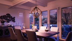 Elegant dining room featuring a wooden table, eight upholstered chairs, a candle centerpiece, and the Urban Ambiance UHP3030 Rustic Chandelier (24-1/2″ x 22″, Olde Iron Finish) from the Wycombe Collection, with large windows framing sunset views of the trees.