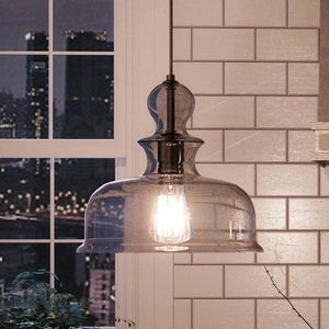 The UHP3041 Modern Farmhouse Pendant Light from Urban Ambiance’s Dundee Collection, featuring an 11-1/4″ x 12″ charcoal-finished frame and exposed bulb, hangs in front of a tiled wall, highlighting its handcrafted artistry while a window reveals city buildings in the background.
