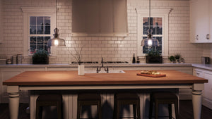 A modern kitchen features white cabinets, a subway tile backsplash, a wooden island with three stools, and two windows overlooking a nighttime cityscape. Overhead, the space is illuminated by pendant lights from the Urban Ambiance Dundee Collection—specifically the UHP3041 Modern Farmhouse Pendant Light with an 11-1/4″ x 12″ smoked glass shade in a charcoal finish.