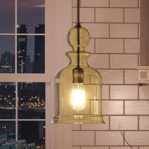 A glowing light bulb inside the Urban Ambiance UHP3045 Modern Farmhouse Pendant Light from the Dundee Collection, featuring a handcrafted glass shade and an Olde Bronze finish, hangs in front of beige subway tile with a cityscape visible through the window.