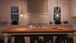 Modern kitchen featuring a white tile backsplash, wooden island countertop, and three stools, illuminated by handcrafted UHP3045 Modern Farmhouse Pendant Lights from Urban Ambiance’s Dundee Collection in an Olde Bronze finish. Two windows reveal a cityscape at dusk, while the space effortlessly blends contemporary design with vintage-modern farmhouse lighting.