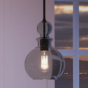 The Urban Ambiance UHP3047 Modern Farmhouse Pendant Light from the Dundee Collection, featuring a handcrafted glass shade and a sleek charcoal finish, hangs illuminated in front of a window at sunset. Outside, clouds and hills are visible, creating a scene that seamlessly blends vintage modern charm with contemporary elegance.