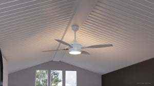 The Matte White UHP9012 Urban Loft Indoor or Outdoor Ceiling Fan from the Mendocino Collection by Urban Ambiance features a built-in light fixture and is installed on a vaulted, white-paneled ceiling. In the background, three windows frame views of trees outside, complementing the fan’s rustic charm.