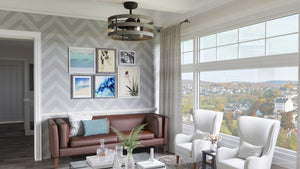A modern living room with a brown sofa, two white chairs, large windows, wall art, and a coffee table with plants is elevated by the Urban Ambiance UHP9031 Modern Farmhouse Indoor Ceiling Fan from the Adelaide Collection (18.1″H x 23.5″W, Charcoal), which adds a touch of rustic industrial style to the space.