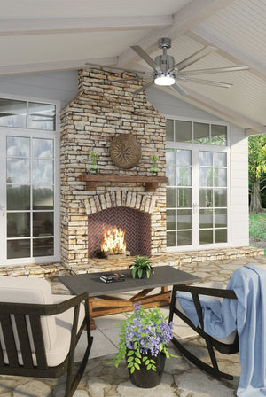 A covered patio features a stone fireplace, two cushioned chairs, a black coffee table with a potted plant, and large windows in the background. Overhead, the Urban Ambiance UHP9051 Urban Loft Indoor / Outdoor Ceiling Fan from the Chatham Collection (16.8″H x 72″W, Brushed Nickel) adds comfort and style with its 8-blade design and integrated LED light while the fire glows in the fireplace.