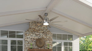 Mounted above a stone fireplace on a covered patio with a white paneled ceiling and large windows, the UHP9051 Urban Loft Indoor / Outdoor Ceiling Fan (16.8″H x 72″W, Brushed Nickel) from the Chatham Collection by Urban Ambiance brings an industrial chic design and integrated LED lighting, adding both stylish function and modern appeal to the space.