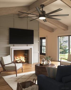 A modern living room featuring the Urban Ambiance UHP9053 Urban Loft Indoor / Outdoor Ceiling Fan from the Chatham Collection in Olde Bronze (16.8″H x 72″W), equipped with a dimmable LED light, along with a fireplace, wall-mounted TV, armchairs, round coffee table, rug, and large windows overlooking trees.