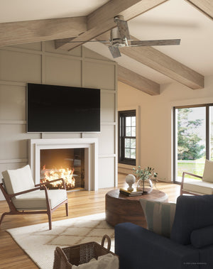 A modern living room features a fireplace, wall-mounted TV, two armchairs, a coffee table, and expansive windows framing lush greenery. The space is enhanced by the Urban Ambiance UHP9061 Urban Loft Indoor Ceiling Fan from the Amelia Collection (15.4″H x 56″W, Aged Nickel), with its reversible blades providing both style and comfort.