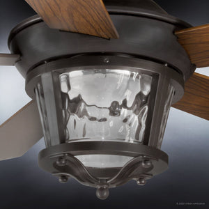 A close-up view of the Urban Ambiance UHP9180 Modern Farmhouse Indoor or Outdoor Ceiling Fan from the Catalina Collection, showcasing its wooden blades and Olde Bronze finish, along with a lantern-style glass light fixture featuring a textured, hammered glass shade. Fan dimensions: 19.5″H x 52″W.