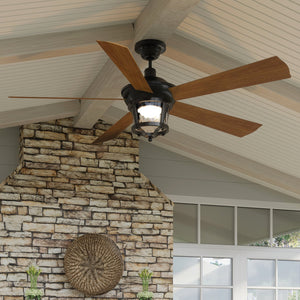 The Urban Ambiance UHP9180 Modern Farmhouse Indoor or Outdoor Ceiling Fan from the Catalina Collection, measuring 19.5 inches high by 52 inches wide and finished in Olde Bronze with wooden blades, features a central light fixture with a hammered glass shade and is installed on a white wood-paneled ceiling above a stone fireplace.
