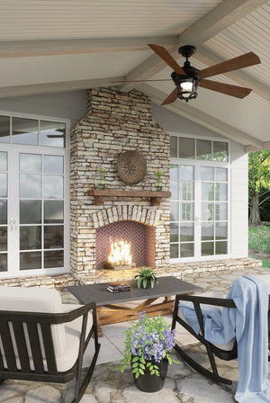 The covered patio features a stone fireplace with a glowing fire, two chairs, and a coffee table adorned with plants. Overhead, an Urban Ambiance UHP9180 Modern Farmhouse Indoor or Outdoor Ceiling Fan from the Catalina Collection in Olde Bronze (19.5″H x 52″W) provides stylish comfort. The space overlooks lush greenery visible through expansive windows and glass doors.