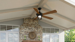 An Urban Ambiance UHP9180 Modern Farmhouse Indoor or Outdoor Ceiling Fan from the Catalina Collection, featuring a 52-inch width, wooden blades, and an Olde Bronze finish, is mounted on a white vaulted ceiling above a stone fireplace and surrounded by windows.
