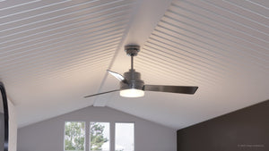 The Urban Ambiance UHP9210 Modern Indoor Ceiling Fan from the Capitola Collection, measuring 15.8 inches high by 52 inches wide with a brushed nickel finish, is mounted on a white vaulted ceiling in a room featuring three windows and dark accent walls. This contemporary ceiling fan effortlessly complements the sleek décor while delivering both efficient airflow and illumination for enhanced style and comfort.