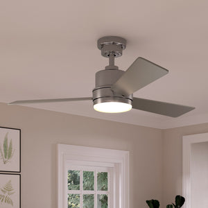 The UHP9221 Modern Indoor Ceiling Fan from Urban Ambiance’s Capitola Collection features a sleek brushed nickel finish, two blades, and a built-in light fixture. Measuring 15.6 inches high by 44 inches wide, this stylish ceiling fan is mounted on a white ceiling in a room decorated with framed artwork and illuminated by natural light from a window.