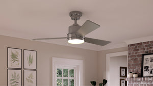 The UHP9221 Modern Indoor Ceiling Fan from Urban Ambiance’s Capitola Collection, featuring a sleek brushed nickel finish, three blades, and an integrated LED light fixture, is installed in a living room adorned with framed art and a brick fireplace.