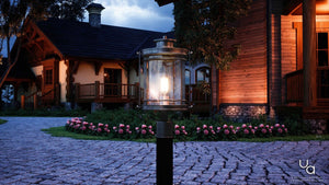 The Urban Ambiance UQL1010 Mid-Century Modern Outdoor Post Light from the Cannes Collection, featuring a black silk finish and measuring 19.5 inches high by 10.5 inches wide, illuminates a cobblestone path near a wooden house at dusk, with blooming flowers and trees in the background.