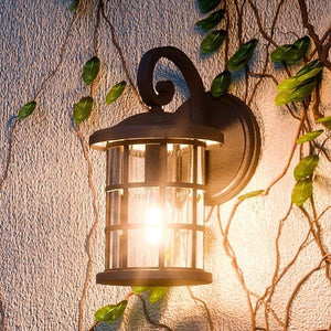 The Urban Ambiance UQL1042 Craftsman Outdoor Wall Light, 14.25"H x 8"W with a natural black finish and marine-grade construction, glows against a textured wall framed by green vines.