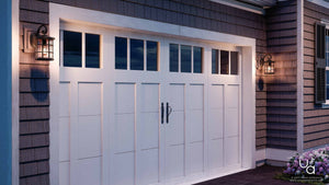 A white paneled garage door with windows at the top is flanked by two Urban Ambiance UQL1043 Craftsman Outdoor Wall Lights from the Vienna Collection, each measuring 14.25″H x 8″W and featuring a Parisian Bronze finish and marine-grade construction. At dusk, the seedy glass of these fixtures emits a warm, inviting glow that enhances the exterior’s charming ambiance.