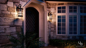 At dusk, the stone house exterior showcases a dark wooden door framed by two illuminated Urban Ambiance UQL1045 Craftsman Outdoor Wall Lights from the Vienna Collection (17.75″H x 10″W), featuring a Parisian Bronze finish and marine-grade construction with seeded glass, set alongside large windows and greenery near the entrance.