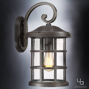 A wall-mounted Urban Ambiance UQL1045 Craftsman Outdoor Wall Light from the Vienna Collection, featuring a marine-grade metal frame in Parisian Bronze finish and seeded glass panels with an exposed bulb, is illuminated against a gray background.