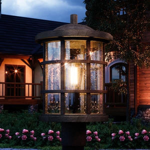 A close-up of the Urban Ambiance UQL1046 Craftsman Outdoor Post Light, 17.25"H x 10"W, Natural Black Finish, Vienna Collection, with marine-grade construction and raindrops on its glass in a garden at dusk with pink flowers and a house behind.