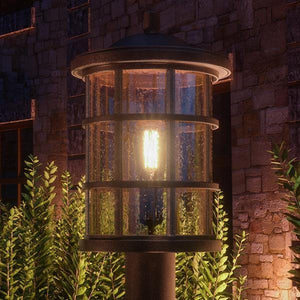 The Urban Ambiance UQL1047 Craftsman Outdoor Post Light, 17.25"H x 10"W, Parisian Bronze from the Vienna Collection glows in front of a stone wall and greenery at night with its marine-grade cylindrical seeded glass design.