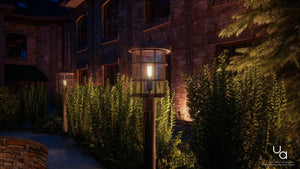 An outdoor pathway illuminated at night with tall Urban Ambiance UQL1047 Craftsman Outdoor Post Lights from the Vienna Collection, featuring a Parisian Bronze finish and marine-grade construction, is bordered by green bushes and set against a stone building exterior.